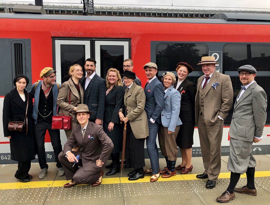 Im Hintergrund steht der rote Kulturzug und am Bahnsteig, direkt vor dem Zug, ist die 20er-Jahre Gruppe zu sehen (13 Personen)