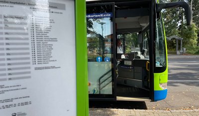 Das Foto zeigt einen Fahrplanaushang an einer Haltestelle und im Hintergrund einen Bus mit geöffneter Vordertür.