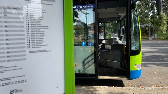 Das Foto zeigt einen Fahrplanaushang an einer Haltestelle und im Hintergrund einen Bus mit geöffneter Vordertür.