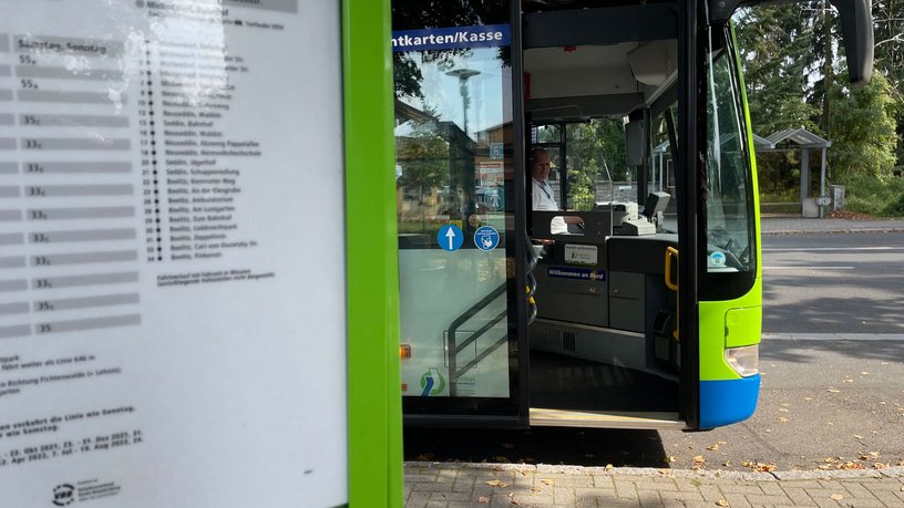 Das Foto zeigt einen Fahrplanaushang an einer Haltestelle und im Hintergrund einen Bus mit geöffneter Vordertür.