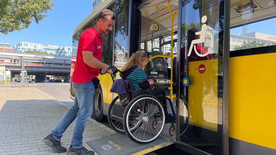 Mitarbeiter des Begleitservices schiebt eine Person im Rollstuhl in einen Bus