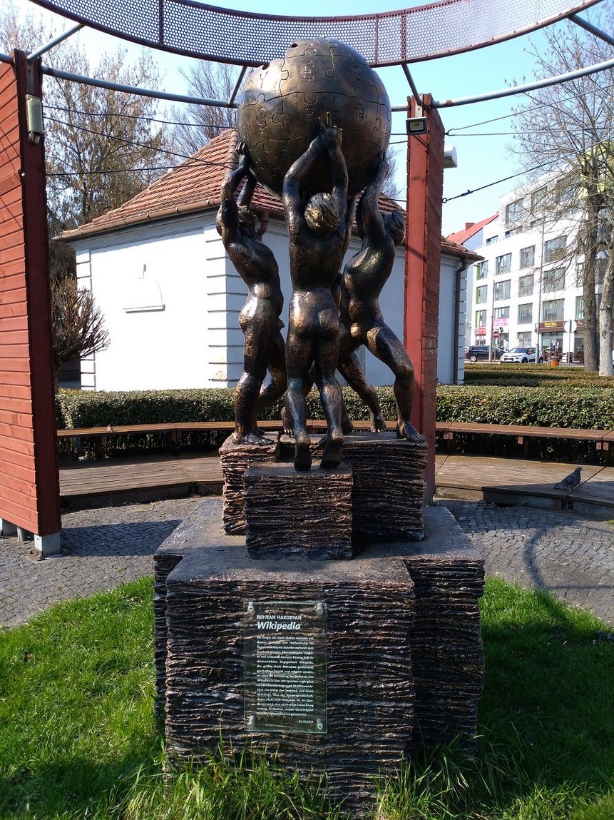 Wikipedia-Denkmal in Słubice. Es hat die Form einer nach oben offenen Weltkugel als Puzzle, also das Logo von Wikipedia, welches von zwei Frauen und zwei Männern getragen wird.