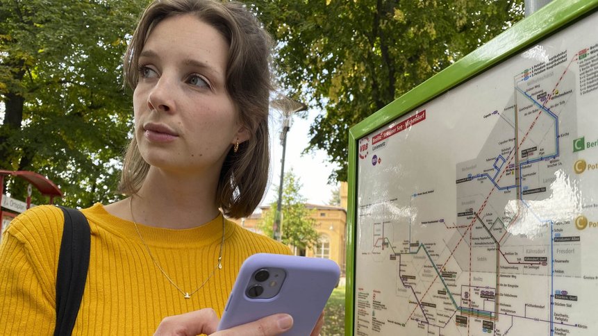 Das Foto zeigt eine junge Frau mit Handy am Bahnhof vor einem Liniennetz.