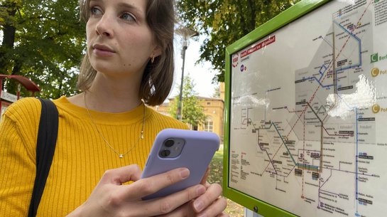 Das Foto zeigt eine junge Frau mit Handy am Bahnhof vor einem Liniennetz.