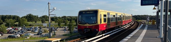 Das Foto zeigt einen Bahnsteig mit S-Bahn sowie im Hintergrund parkende Autos.