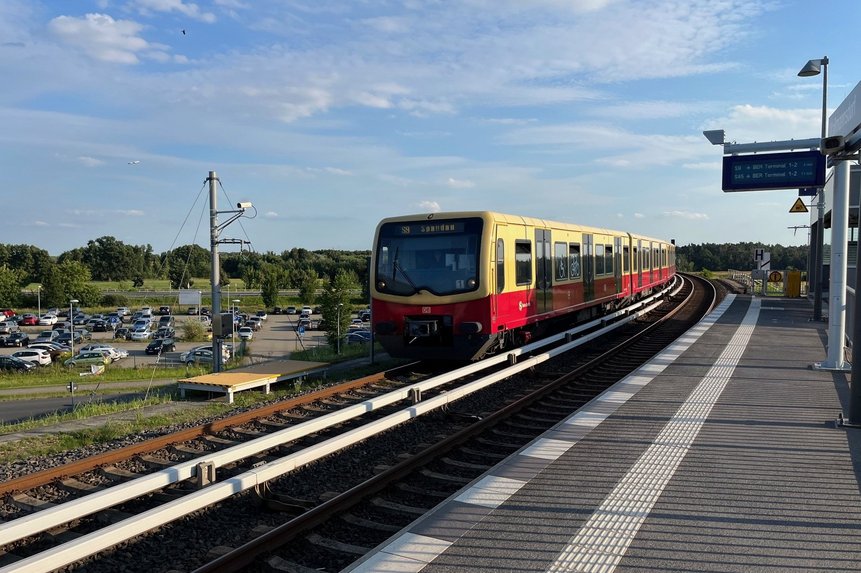 Park & Ride am S-Bahnhof Waßmannsdorf. Das Foto zeigt einen Bahnsteig mit S-Bahn sowie im Hintergrund parkende Autos.