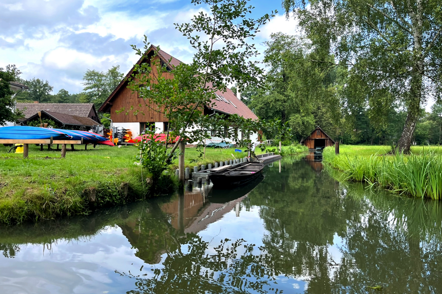 Das Foto zeigt Wasser, Wiese und ein Haus mit Holzdach sowie daneben Kanus und einen Spreewaldkahn.