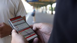 Zwei Personen am Bahnsteig. Eine davon hält ein Smartphone mit der geöffneten App.